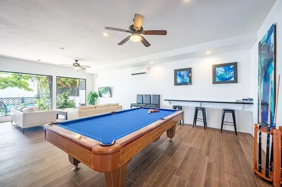 fotografia-para-propiedades-espacios-28 | Pacific Photography Mesa de billar con fieltro azul en una luminosa sala de juegos, suelos de madera, ventiladores de techo y una zona de asientos junto a puertas de cristal con vista a un patio exterior.