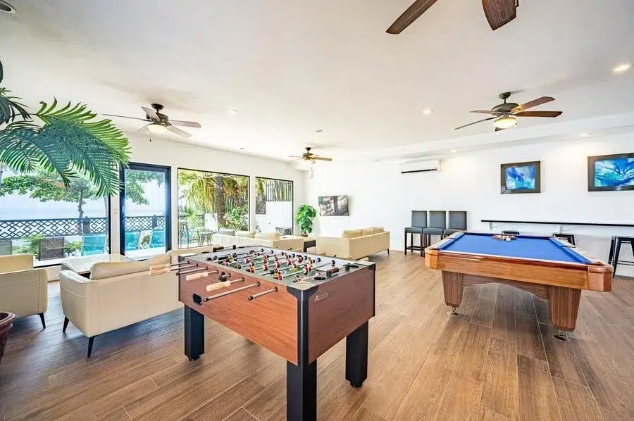 fotografia-para-propiedades-espacios-29 | Pacific Photography Luminosa sala de juegos con una mesa de futbolín y una mesa de billar azul, sofás beige y grandes puertas corredizas que dan a una zona de piscina exterior.