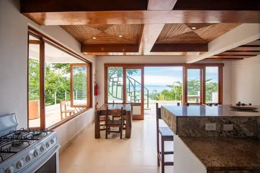fotografia-para-propiedades-espacios-3 | Pacific Photography Cocina y comedor luminosos con una isla de granito, vigas de madera y grandes ventanas que se abren a un patio con vista al mar.