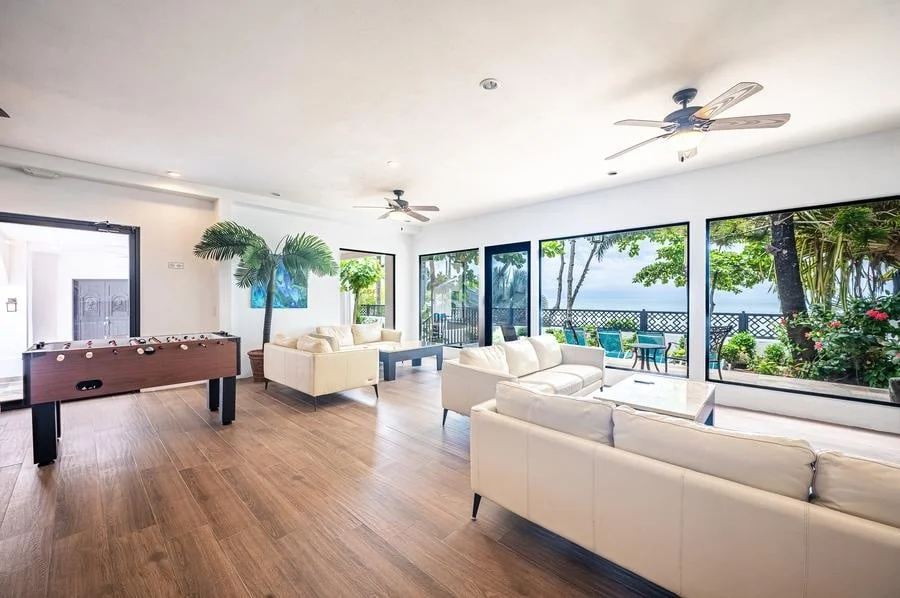 fotografia-para-propiedades-espacios-30 | Pacific Photography Moderna sala de estar luminosa con sofás blancos, suelos de madera y ventanales de suelo a techo que muestran un patio junto al mar y palmeras.