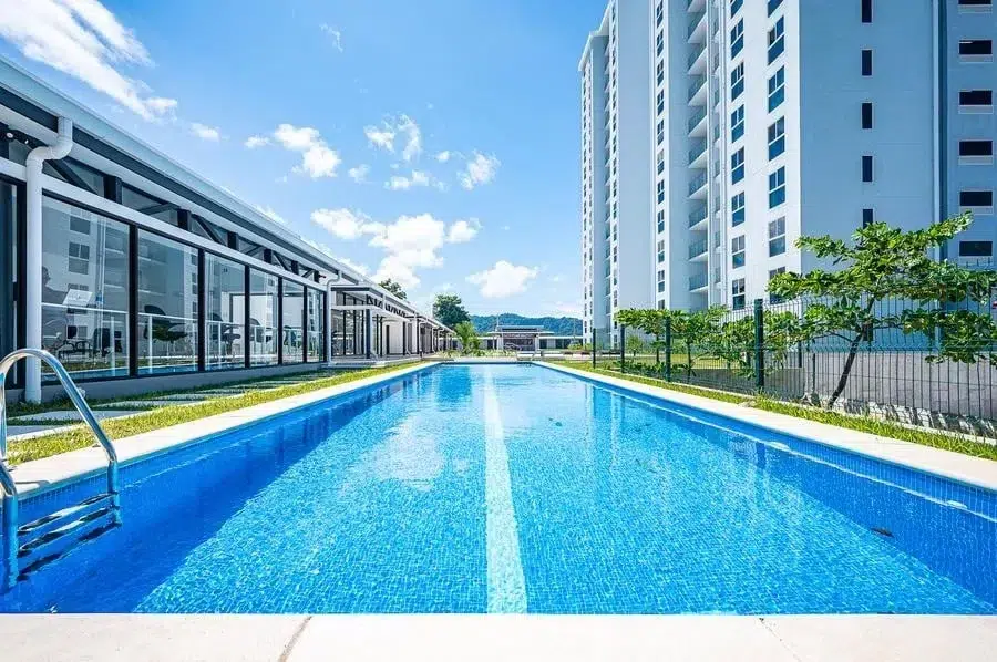 fotografia-para-propiedades-espacios-35 | Pacific Photography Piscina azul rectangular en un moderno complejo residencial con edificios con frente de vidrio y un alto bloque de apartamentos blanco bajo un cielo soleado.