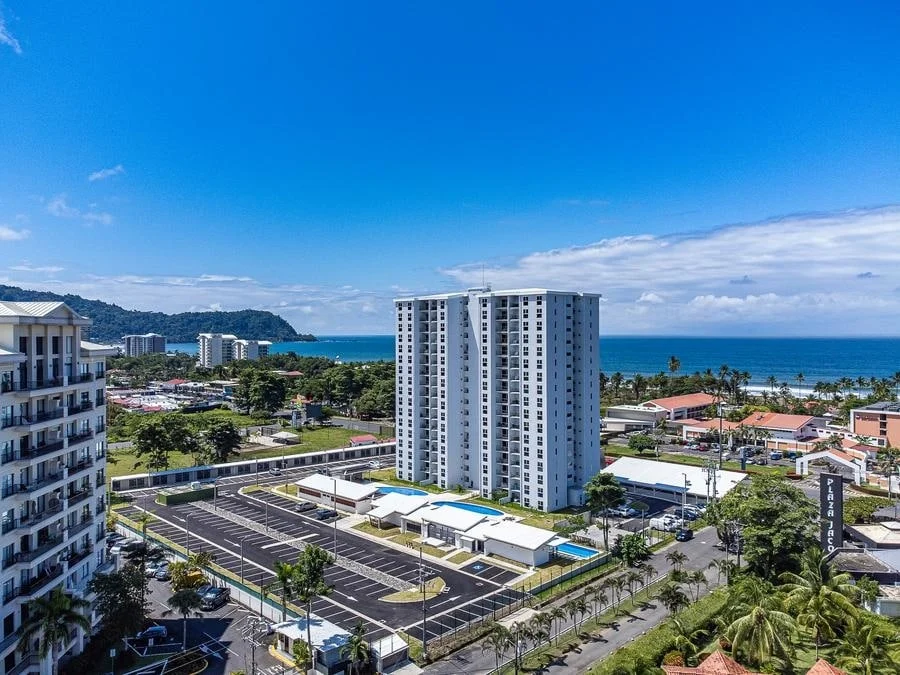 fotografia-para-propiedades-espacios-36 | Pacific Photography Vista aérea de un edificio de apartamentos blanco de gran altura junto a la playa, con una zona de piscina azul, estacionamientos circundantes y palmeras cercanas.