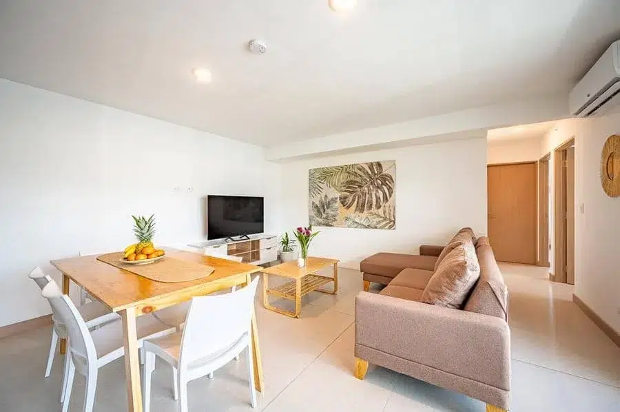 fotografia-para-propiedades-espacios-38 | Pacific Photography Sala de estar y comedor de planta abierta: mesa de comedor de madera con sillas blancas, sofá beige, TV montada en la consola, arte de pared tropical.