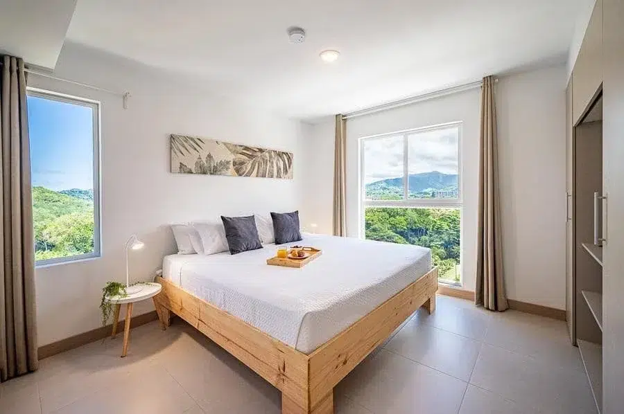 fotografia-para-propiedades-espacios-39 | Pacific Photography Luminoso dormitorio de hotel con una cama de plataforma de madera, ropa de cama blanca y dos cortinas neutras; una pequeña mesa redonda con una lámpara y una planta se encuentra junto a la cama, y una bandeja con el desayuno descansa sobre el edredón mientras grandes ventanas revelan colinas verdes en el exterior.
