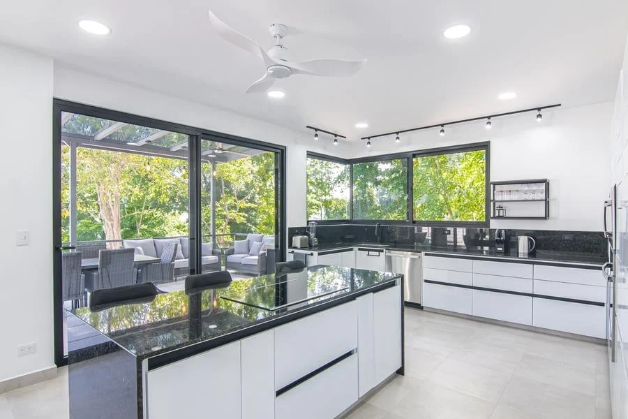 fotografia-para-propiedades-espacios-41 | Pacific Photography Cocina moderna y luminosa con gabinetes blancos y encimeras de granito negro, grandes ventanales y una isla con vistas a una zona de asientos en el patio.
