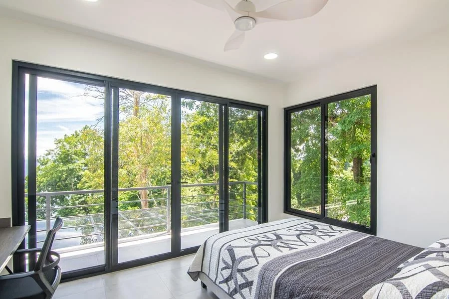 fotografia-para-propiedades-espacios-43 | Pacific Photography Dormitorio luminoso con grandes puertas corredizas de vidrio con marco negro que dan a un balcón y una ventana, mostrando árboles verdes en el exterior; cama con una manta gris estampada visible.