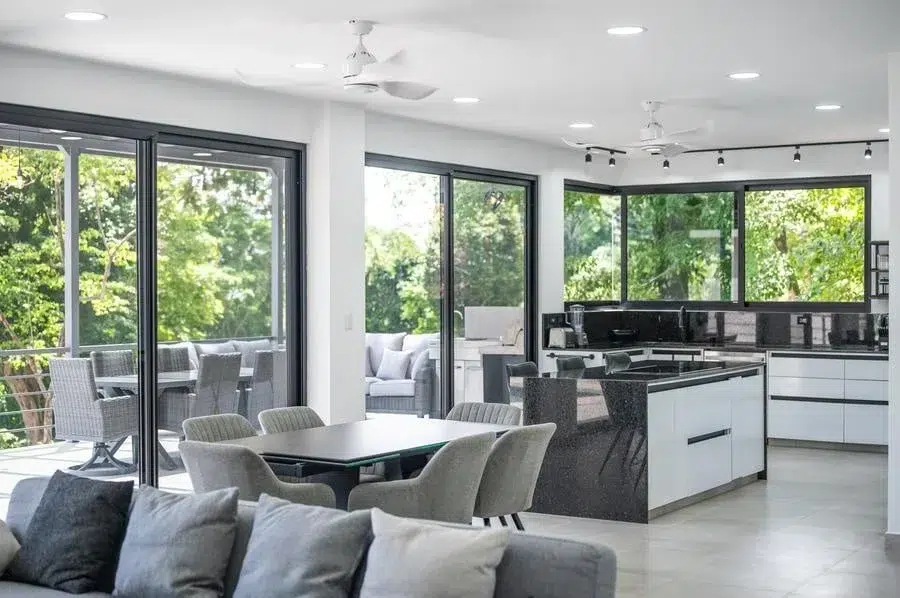 fotografia-para-propiedades-espacios-44 | Pacific Photography Luminosa cocina y comedor de planta abierta con grandes puertas de vidrio que se abren a un patio verde exterior; sillas de comedor grises alrededor de una mesa oscura y una isla con encimera negra.