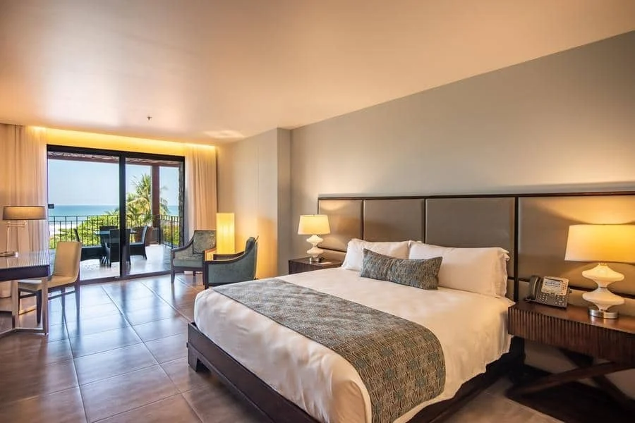fotografia-para-propiedades-espacios-9 | Pacific Photography Habitación de hotel con una cama king, mesitas de noche y vista al balcón del océano y palmeras fuera de las puertas de cristal.