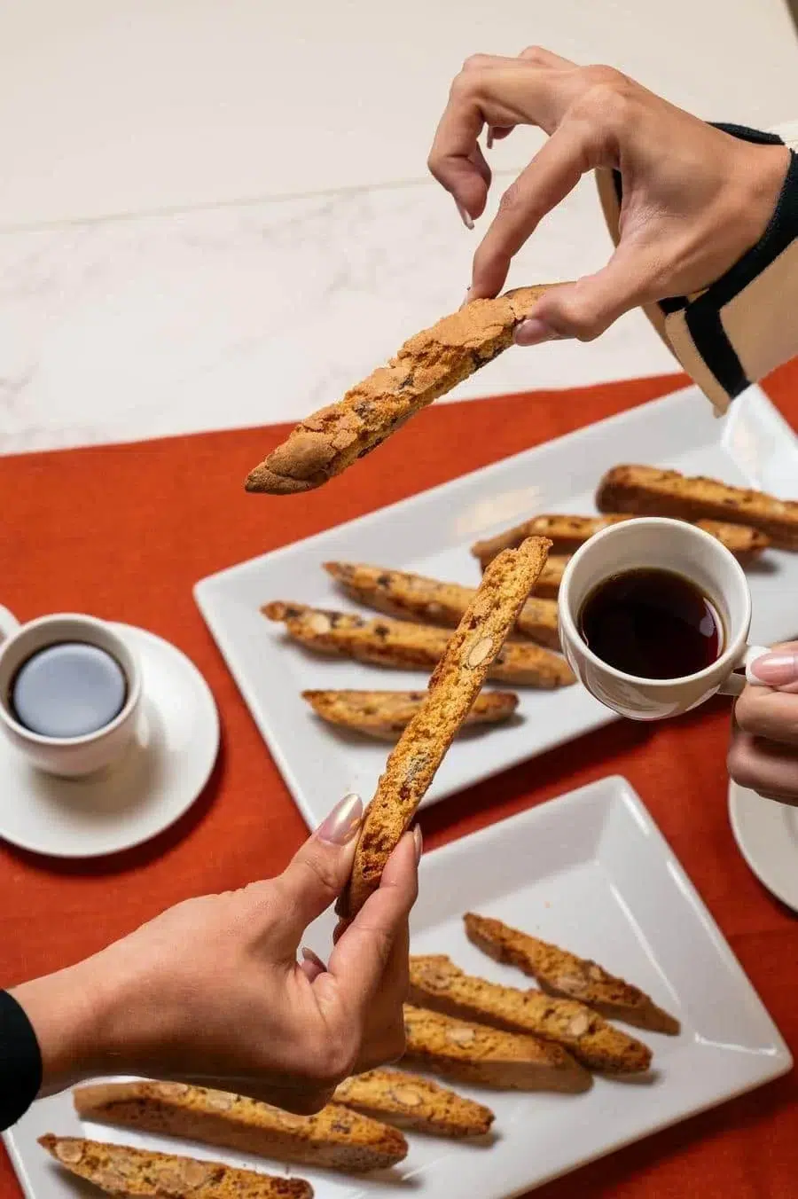 People share biscotti pieces over a white plate and a cup of coffee on a red table.