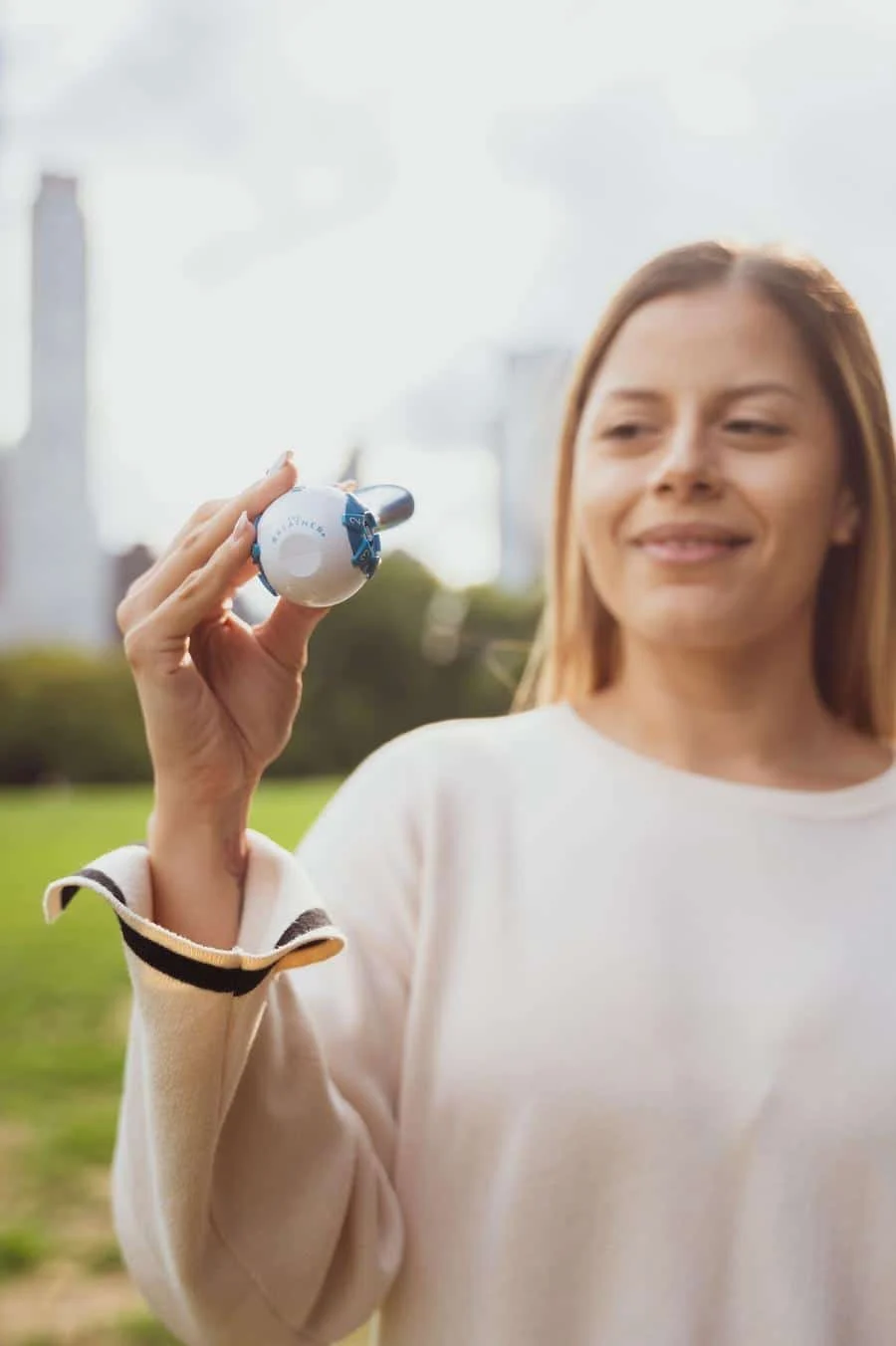 product photography costa rica -7 | Pacific Photography Smiling woman outdoors holding a white golf ball toward the camera, with a blurred city skyline in the background.