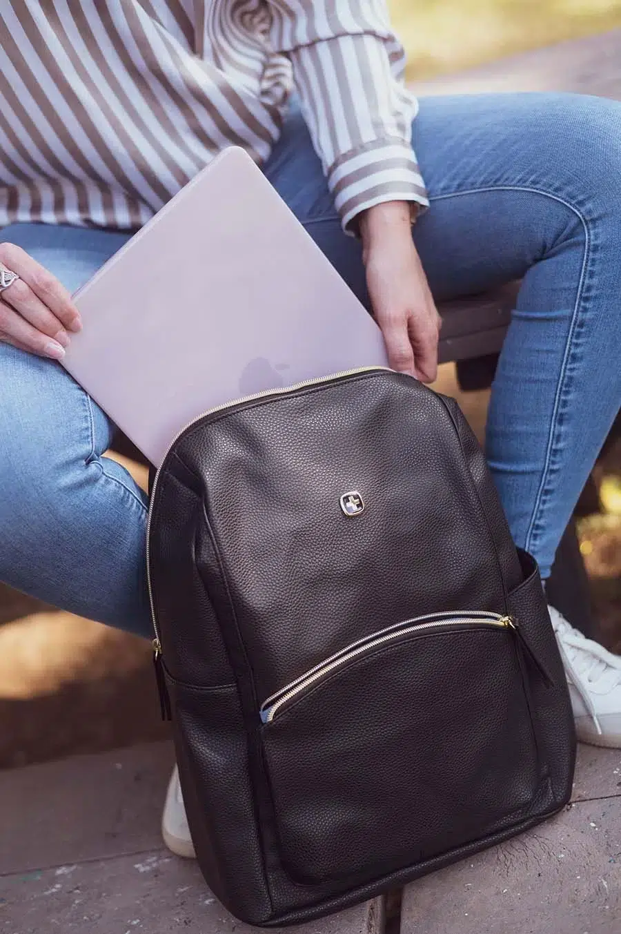 Person in a striped shirt sits outdoors with a black backpack, placing a MacBook into the main compartment of his backpack.