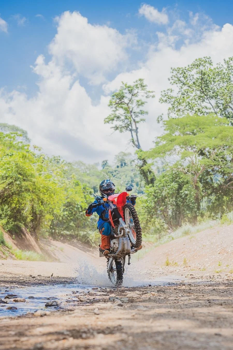 sports and adventure photography costa rica -14 | Pacific Photography Motocross rider on a red dirt bike performs a wheelie across a muddy trail with trees and blue sky in the background.