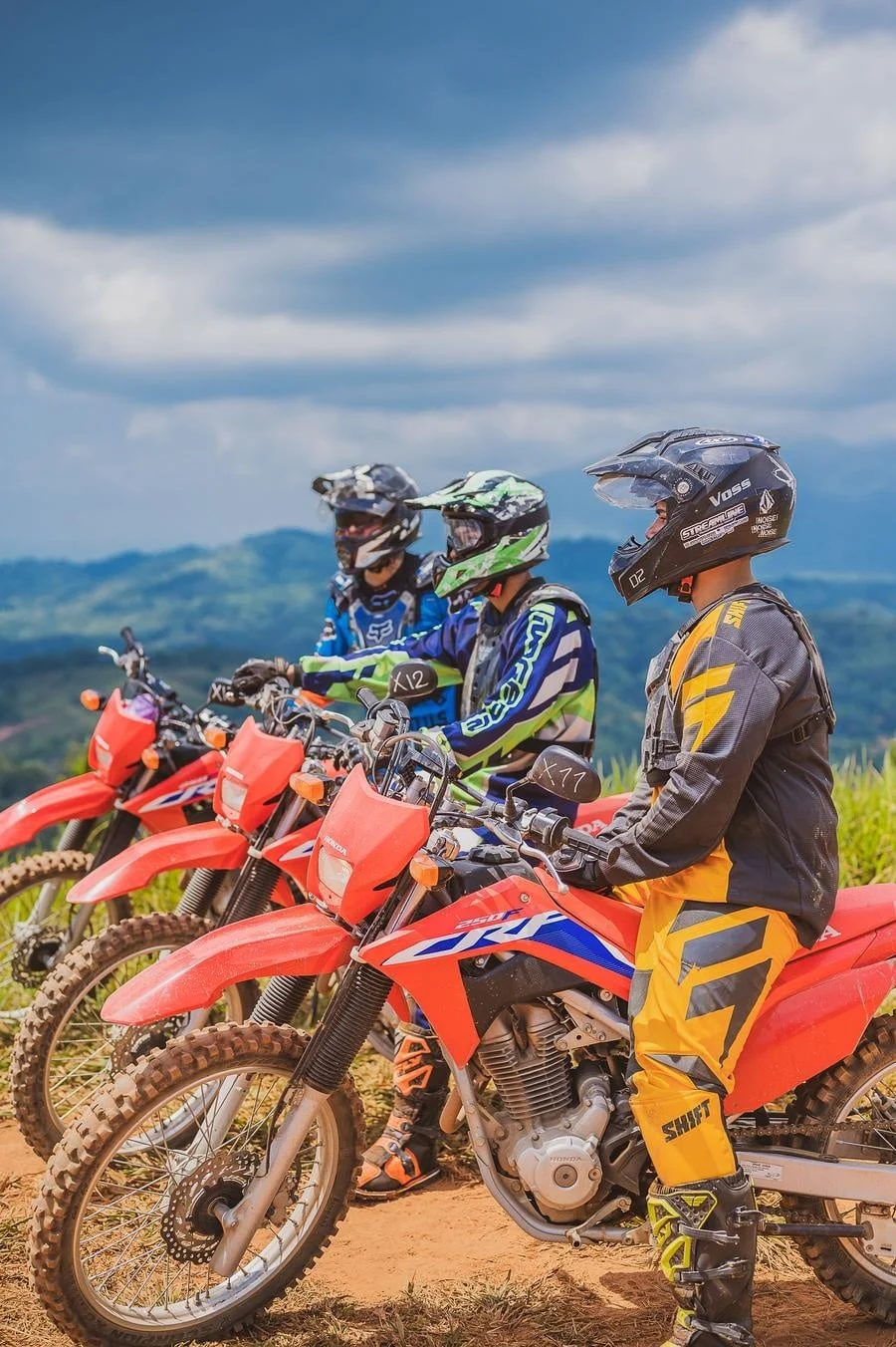 Three motocross riders on red dirt bikes wearing helmets and colorful gear, lined up on a dirt track against a scenic hilly landscape.