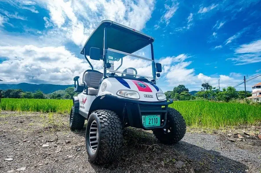 sports and adventure photography costa rica -3 | Pacific Photography White EZGO golf cart with oversized off-road tires parked on a dirt path in a green field under a blue sky and scattered clouds.