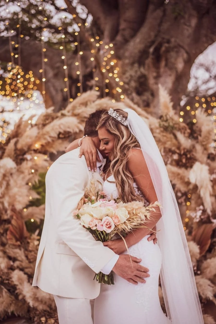 wedding ceremony photography costa rica-1 | Pacific Photography Bride and groom embrace at a wedding, bouquet of pink roses and pampas grass in foreground, warm fairy lights in the background.