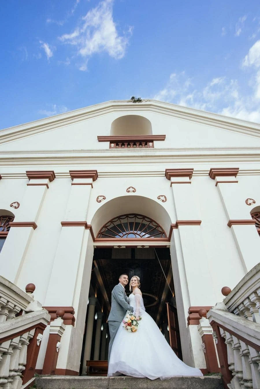 wedding ceremony photography costa rica-10 | Pacific Photography Bride and groom pose at the church entrance on stone steps, the bride holding a bouquet and wearing a white wedding gown with a long train.