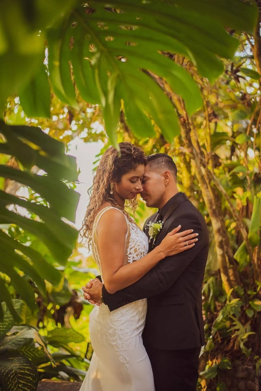 wedding ceremony photography costa rica-11 | Pacific Photography A bride in a lace white wedding gown and a groom in a black suit share a tender embrace in a lush tropical garden with warm sunlight.