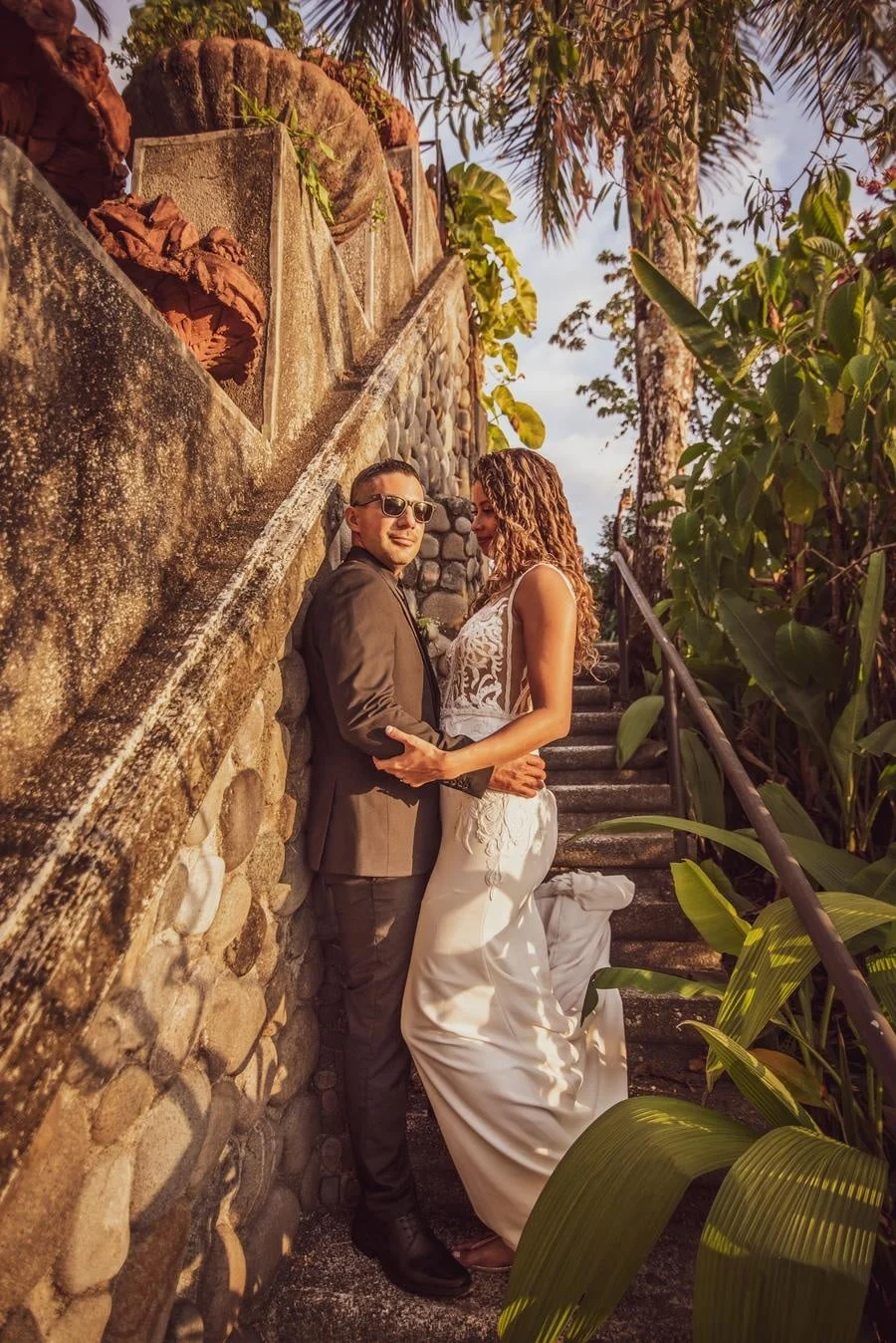 wedding ceremony photography costa rica-12 | Pacific Photography Bride in a lace-illusion white gown and groom in a brown suit embrace on a stone stairway, surrounded by tropical plants and palm trees on a sunny day.