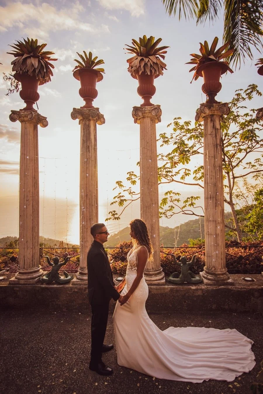 wedding ceremony photography costa rica-13 | Pacific Photography Bride in a white gown and groom in a dark suit hold hands in front of four tall stone columns at sunset.