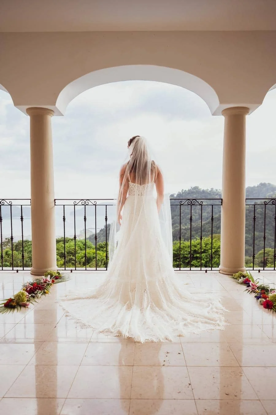 wedding ceremony photography costa rica-14 | Pacific Photography Bride in a white wedding gown with a long train and veil stands on a balcony, facing a scenic hill landscape.