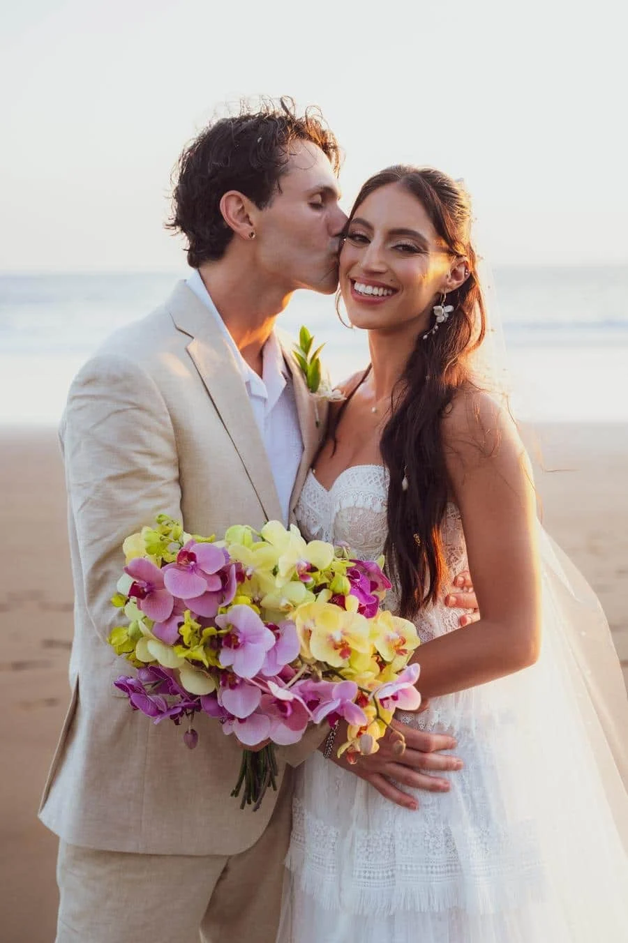 wedding ceremony photography costa rica-18 | Pacific Photography Couple on the beach in wedding attire; the groom kisses the bride on the cheek as she smiles, holding a colorful orchid bouquet.