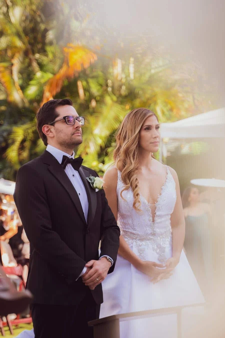 wedding ceremony photography costa rica-19 | Pacific Photography A bride in a lace white wedding dress and a groom in a black tuxedo stand side by side outdoors, awaiting the ceremony with greenery in the background.