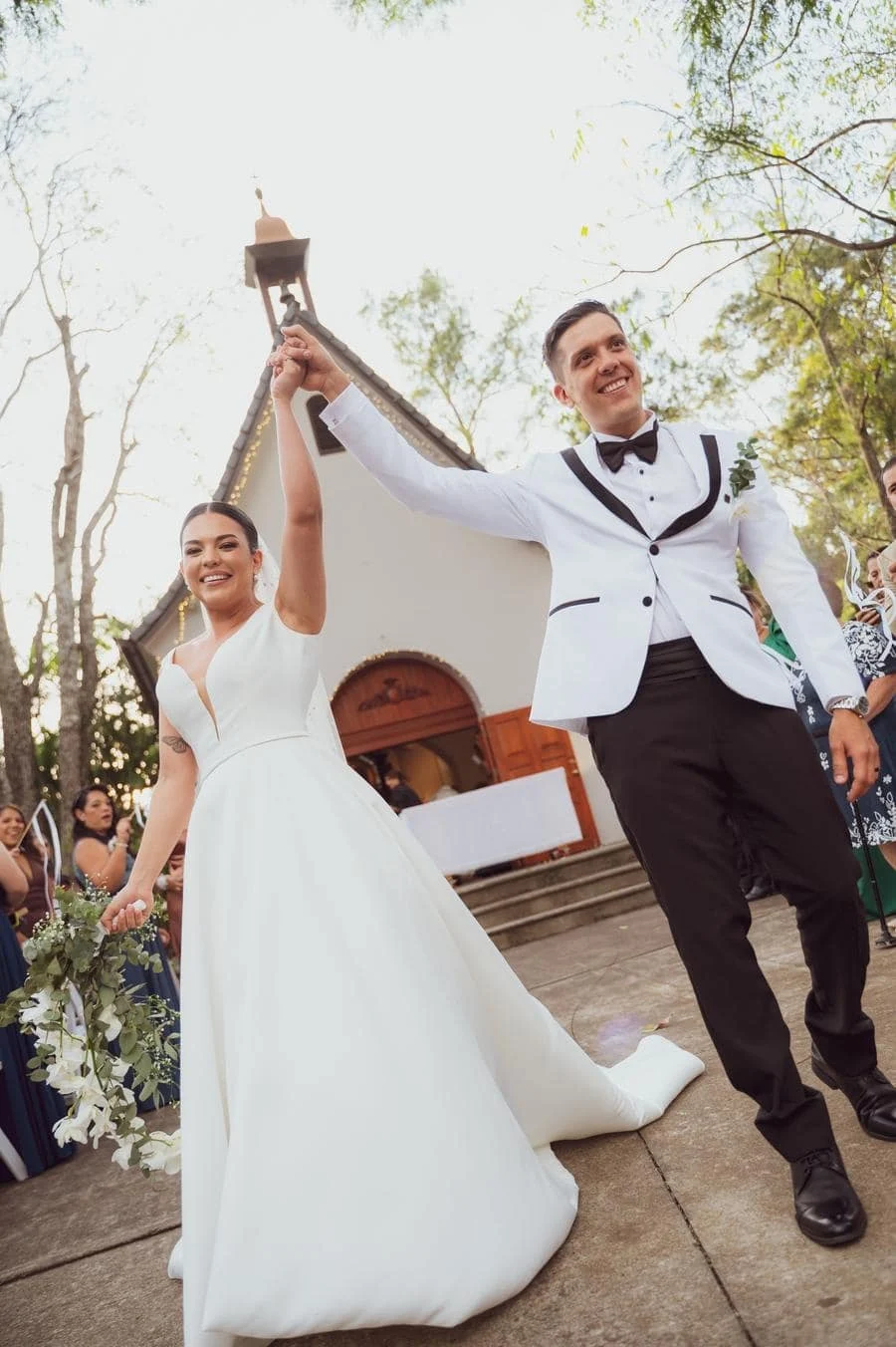 wedding ceremony photography costa rica-20 | Pacific Photography Bride and groom celebrate exiting a chapel, the bride in a white gown holding a bouquet while the groom in a white suit raises their joined hands.
