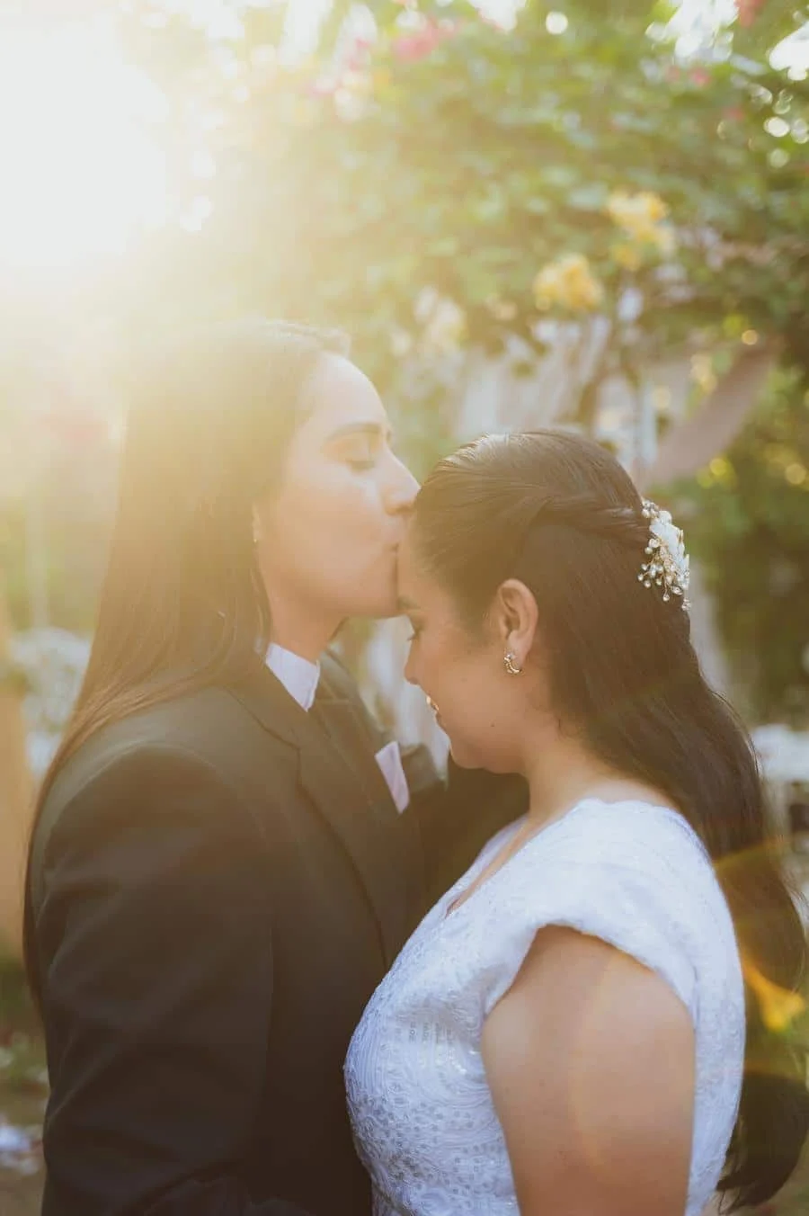 wedding ceremony photography costa rica-23 | Pacific Photography Outdoor wedding moment: one person in a dark suit kisses the forehead of another in a white lace dress as warm sunlight glows between them.
