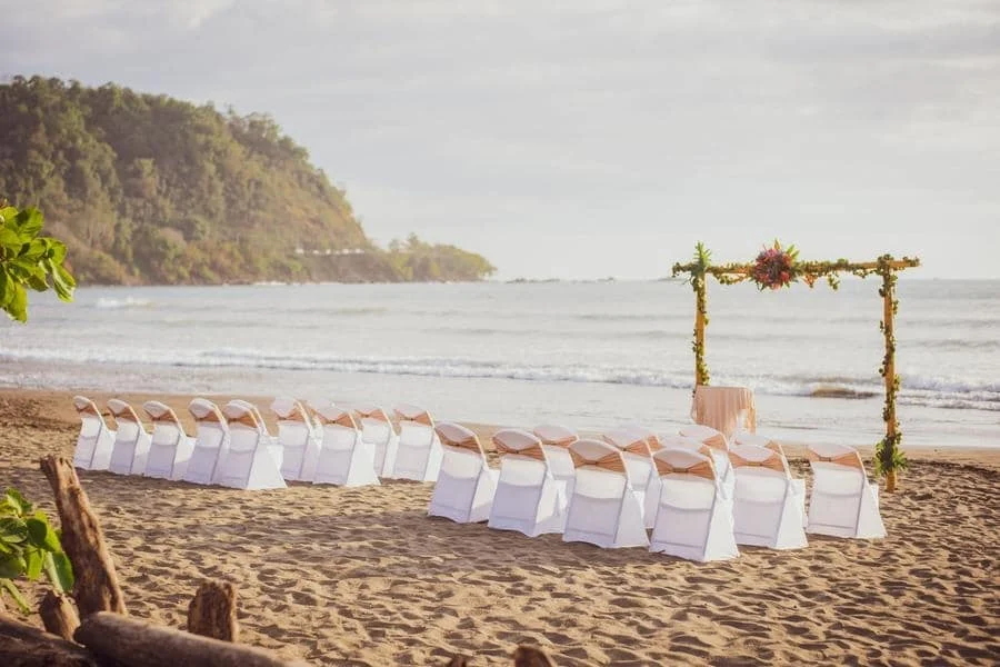 wedding ceremony photography costa rica-4 | Pacific Photography Beach wedding setup on the sand with white chairs in rows facing a flower-adorned arch by the sea.