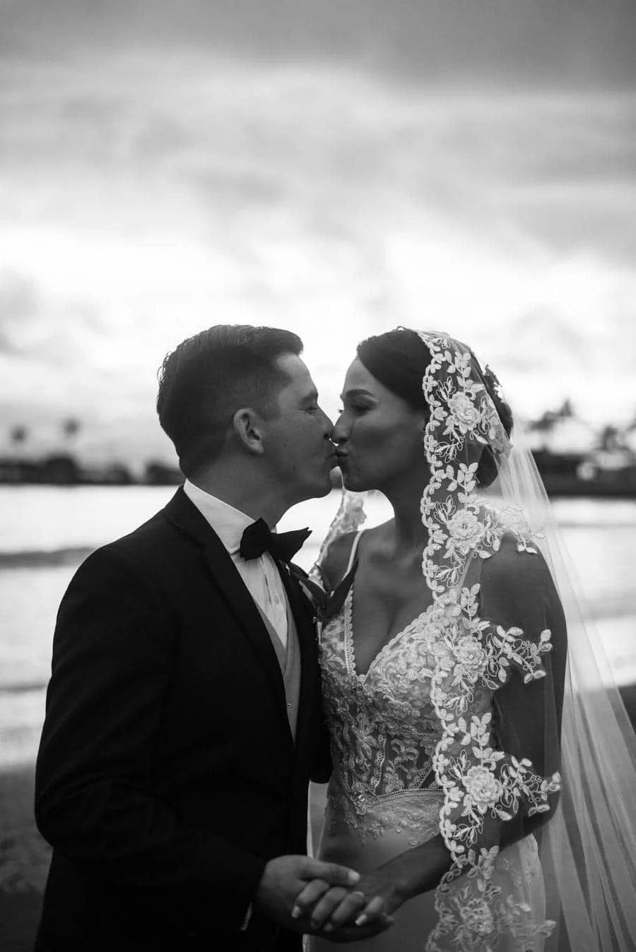 wedding ceremony photography costa rica-5 | Pacific Photography Black-and-white photo of a bride and groom kissing by the water; bride wears an intricate lace veil and gown, groom in a suit with a bow tie.