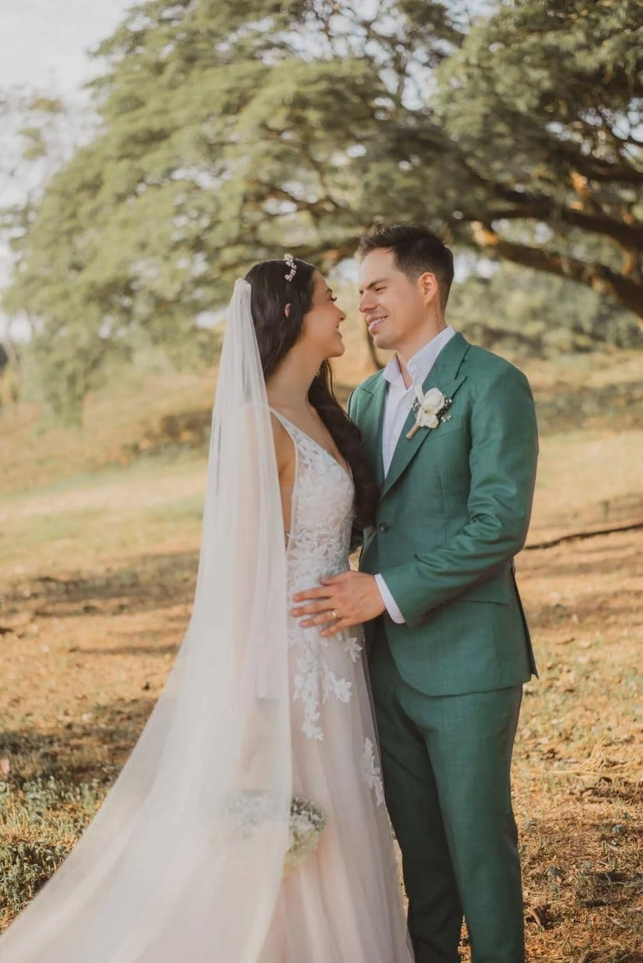 wedding ceremony photography costa rica-7 | Pacific Photography Bride in a lace gown and veil embraces groom in a green suit outdoors, smiling at each other beneath trees at a sunny wedding.