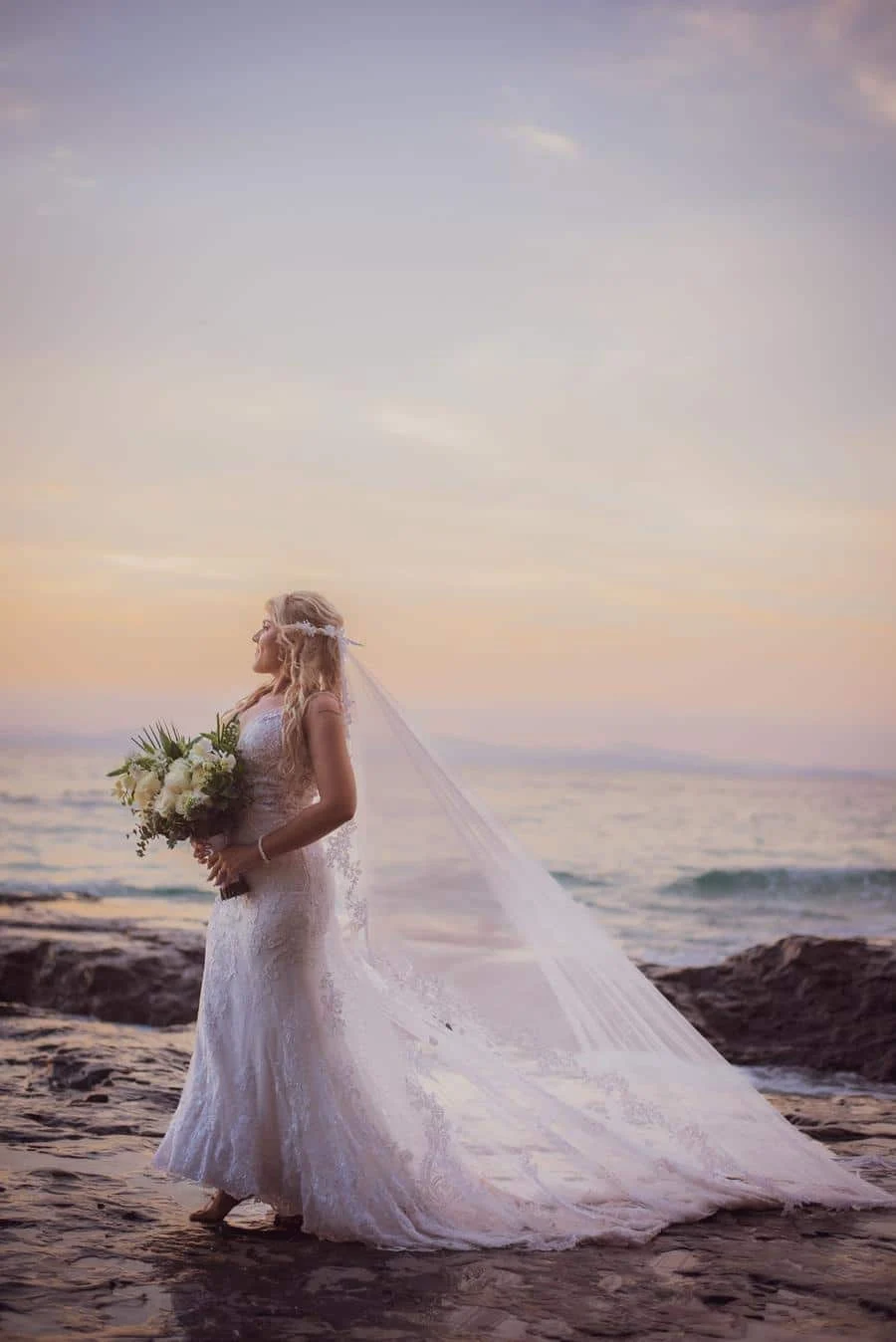 wedding ceremony photography costa rica-9 | Pacific Photography Bride in a lace wedding gown on a rocky beach at sunset, holding a bouquet of white flowers.