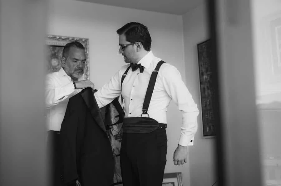 wedding prep photography costa rica-10 | Pacific Photography Two men in formal wear; one helps the other put on a suit jacket in a doorway, both dressed in white shirts and bow ties.