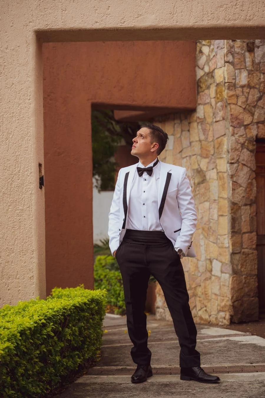 wedding prep photography costa rica-12 | Pacific Photography Man in a white tuxedo jacket, black bow tie, and black pants stands outdoors beside a stone wall, hands in pockets, looking up.