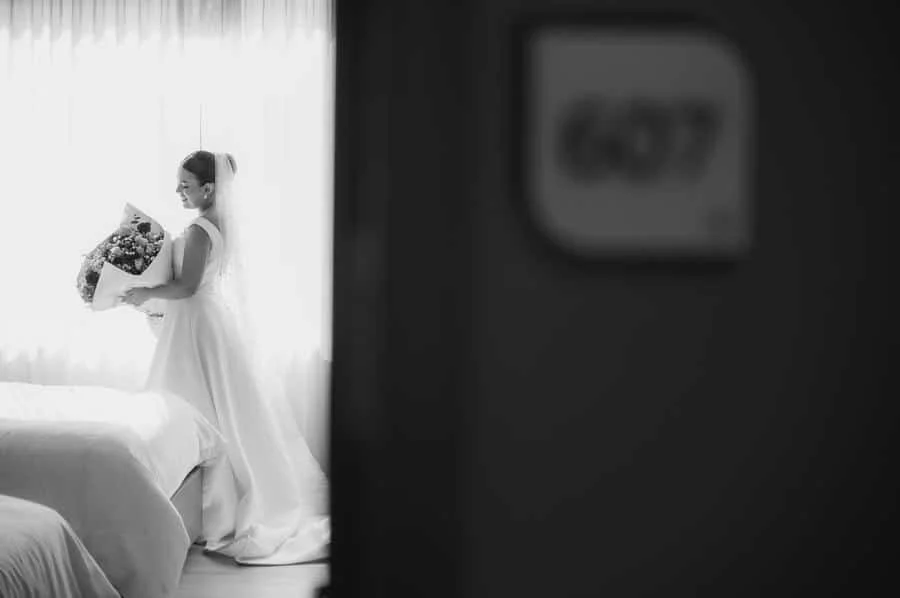 wedding prep photography costa rica-14 | Pacific Photography Bride in a white wedding gown holding a bouquet, standing by a sunlit window, viewed from a doorway in a dim foreground.