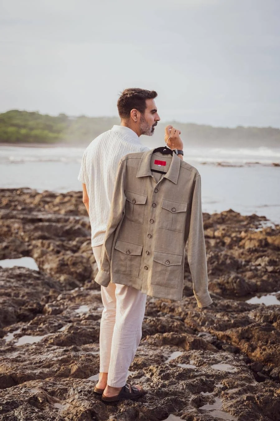 wedding prep photography costa rica-17 | Pacific Photography Man standing on a rocky beach holding a beige jacket on a hanger, gazing toward the sea.