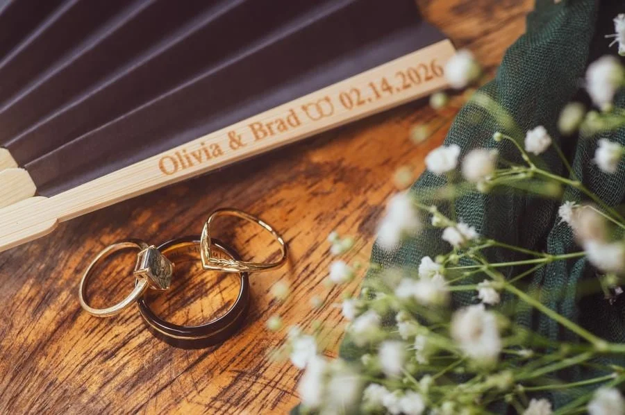 wedding prep photography costa rica-18 | Pacific Photography Engraved wooden keepsake reading 'Olivia & Brad 02.14.2026' with two rings and a floral bouquet on a wooden table behind.