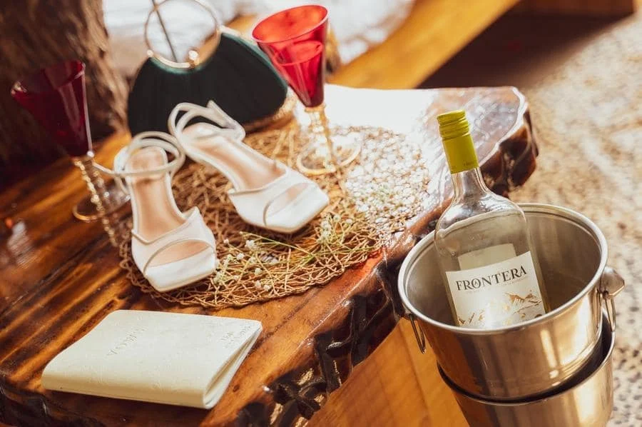 wedding prep photography costa rica-19 | Pacific Photography Wedding table setup with white high-heeled shoes, a guest book, and a bottle of wine chilling in a metal bucket on a rustic wooden surface.