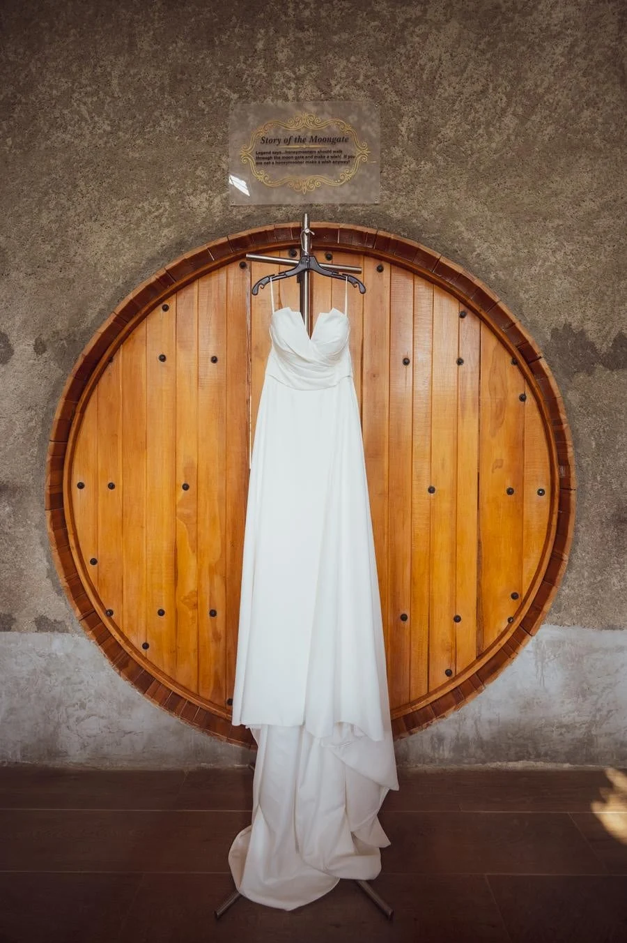 wedding prep photography costa rica-20 | Pacific Photography White wedding dress on a hanger in front of a large round wooden door with a sign above that reads 'Story of the Moongate'.