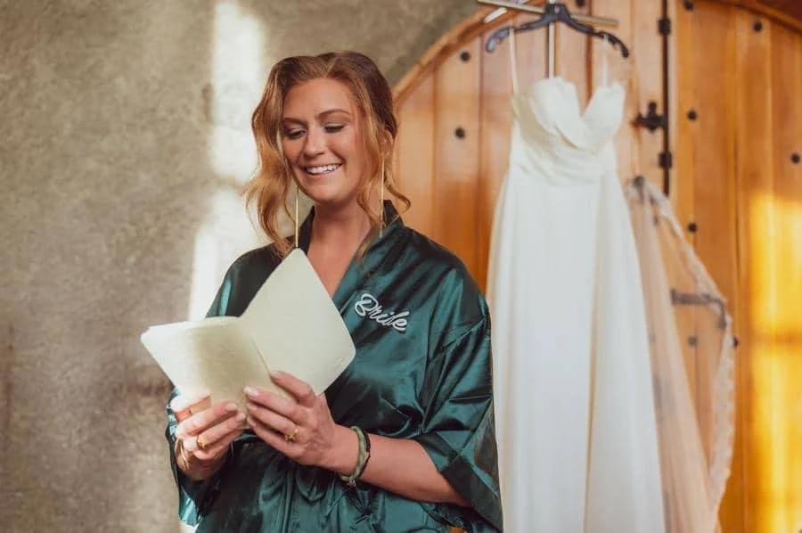 wedding prep photography costa rica-22 | Pacific Photography Smiling bride-to-be in a green satin robe reads a card, with a white wedding dress hanging on a rack behind her in a rustic room.