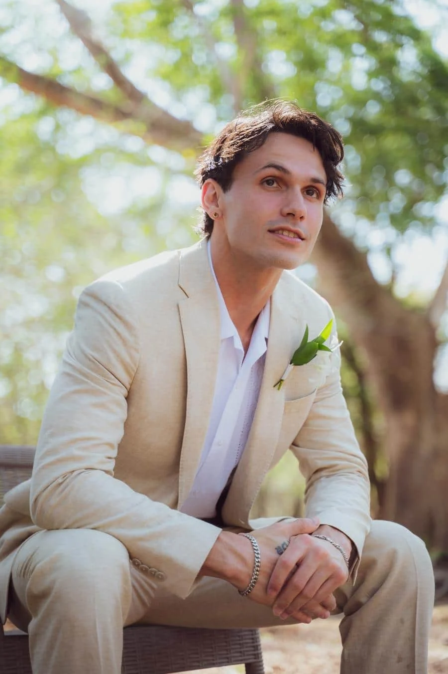 wedding prep photography costa rica-26 | Pacific Photography Man in a light beige suit with a white shirt and boutonniere sits on a bench outdoors, hands clasped.
