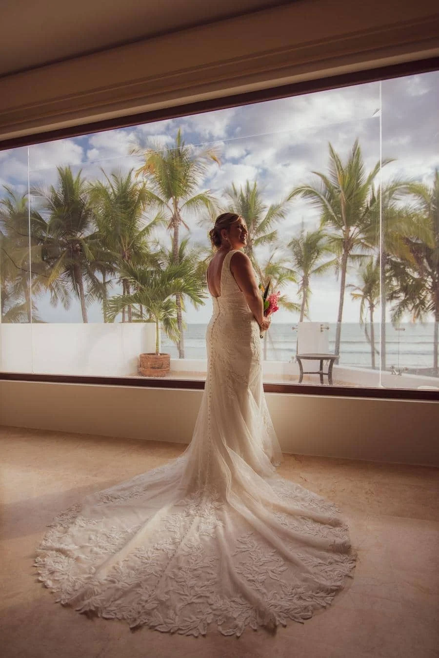 wedding prep photography costa rica-4 | Pacific Photography Bride in a lace wedding gown with a long train standing indoors by a large window, looking toward the ocean and palm trees outside.