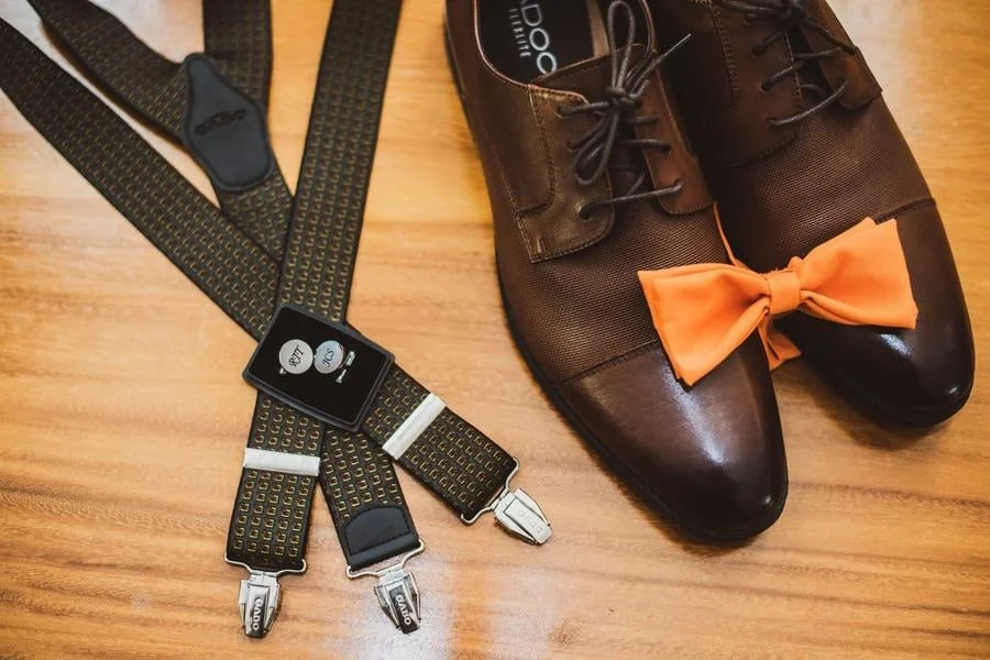 wedding prep photography costa rica-5 | Pacific Photography Brown dress shoes with an orange bow on the toe sit beside black patterned suspenders and a small smartwatch on a wooden floor, arranged as a formal outfit.