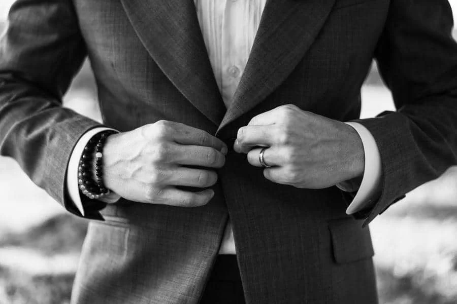 wedding prep photography costa rica-6 | Pacific Photography Close-up of a person in a suit buttoning the blazer, wearing a ring and beaded bracelet on the left wrist (black-and-white photo).