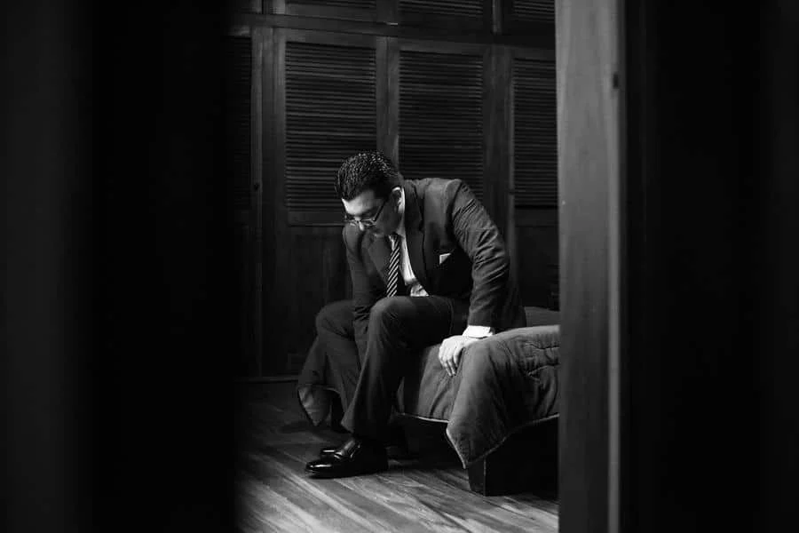 wedding prep photography costa rica-9 | Pacific Photography Black-and-white photo of a man in a suit sitting on the edge of a sofa, head bowed, in a dim wood-paneled room.