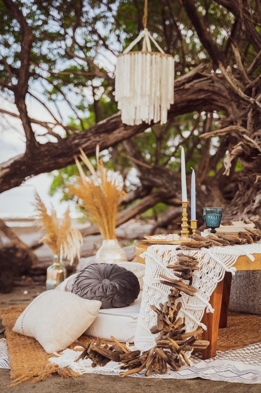 wedding reception and party photography in costa rica -10 | Pacific Photography Boho outdoor beach setup with a lace table runner, candles, cushions, and driftwood decor under a tree.