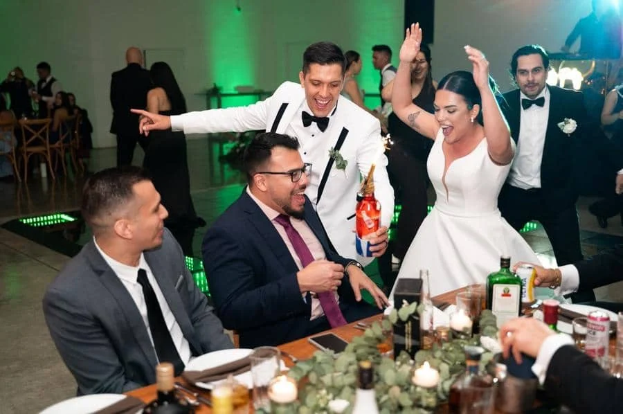 wedding reception and party photography in costa rica -11 | Pacific Photography Bride in a white dress dances with arms raised as guests toast at a wedding reception with a decorated table in the foreground.