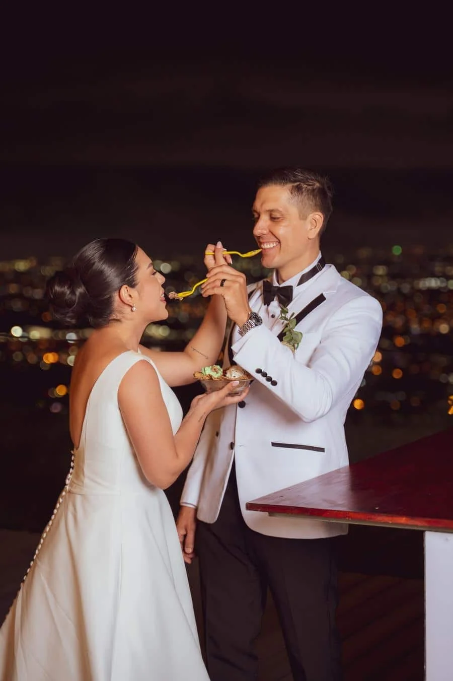 wedding reception and party photography in costa rica -12 | Pacific Photography Bride and groom in wedding attire share a bite of pasta on a rooftop at night, city lights in the background.