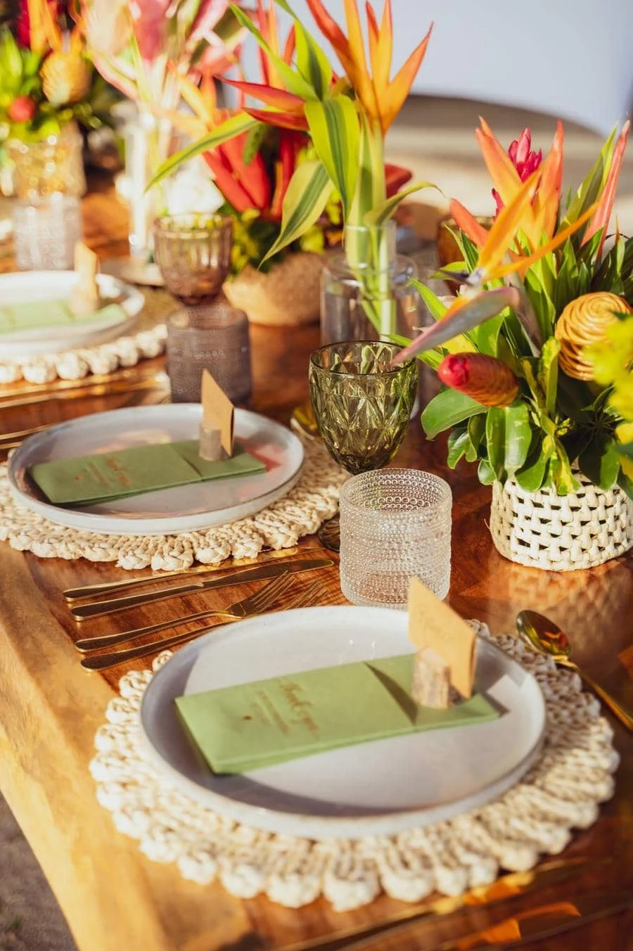 wedding reception and party photography in costa rica -6 | Pacific Photography Table set for a formal meal with green napkins on white plates, gold flatware, and a vibrant tropical floral centerpiece in woven pots.