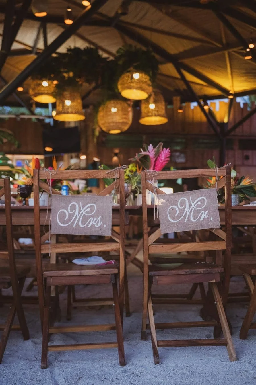 wedding reception and party photography in costa rica -7 | Pacific Photography Rustic wedding reception setup with wooden chairs labeled ‘Mrs.’ and ‘Mr.’ hanging on the back, under warm hanging lanterns.
